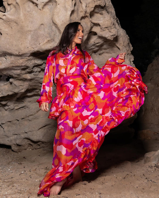 Smiling woman in vibrant pink, orange, and red floral maxi dress dancing barefoot on sandy ground near rocky formations