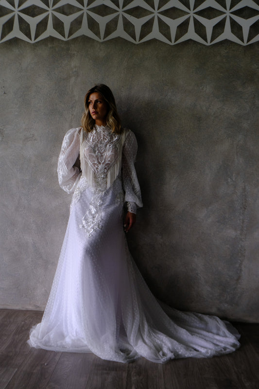 Woman in detailed white lace wedding gown with puff sleeves standing against textured gray wall
