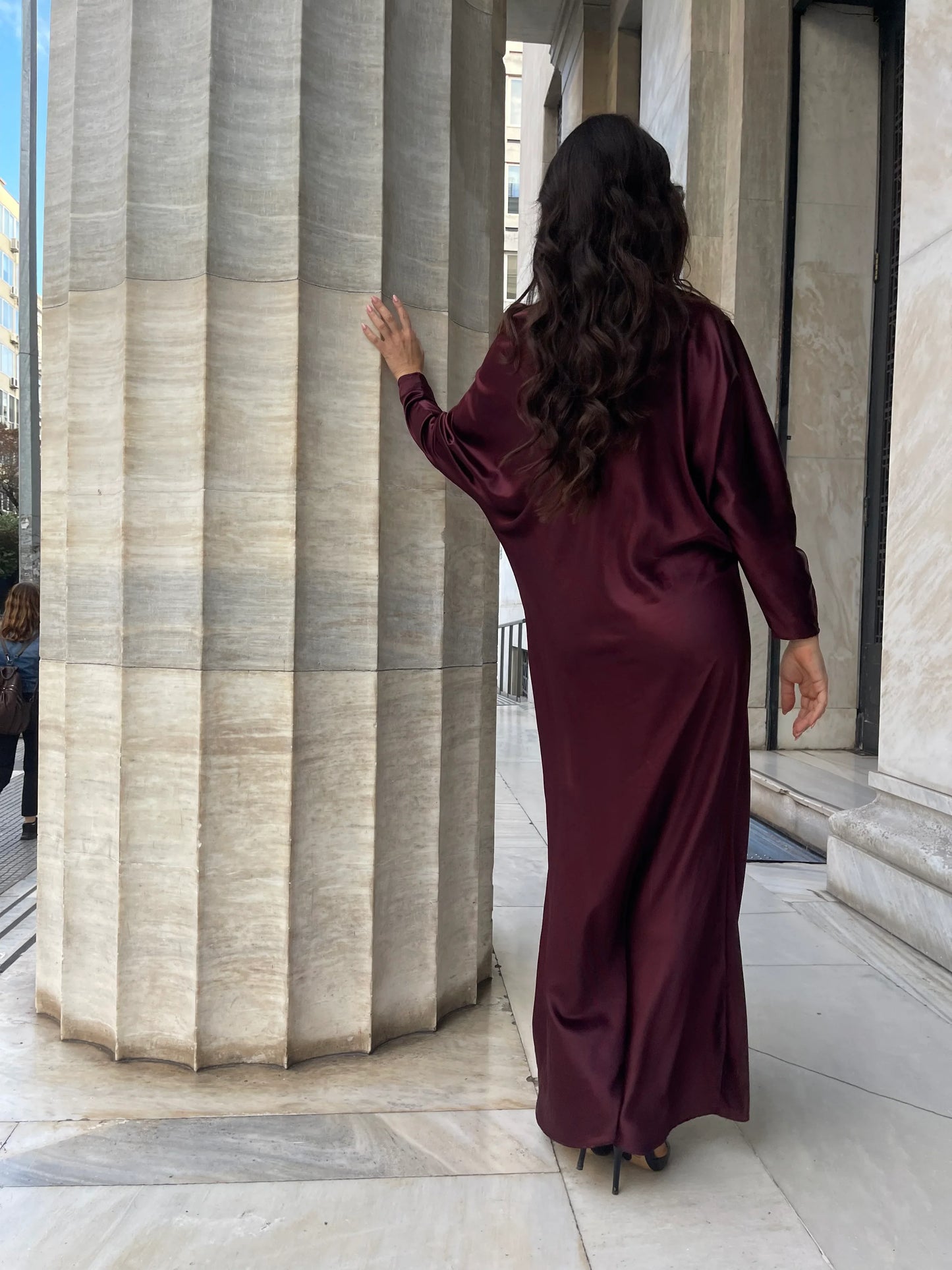 Woman in long burgundy satin dress with heels, standing by large marble column outdoors