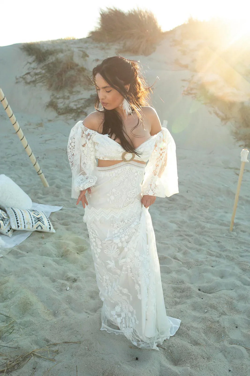 Woman in white lace off-shoulder dress standing on sandy beach with sunlight and boho decor