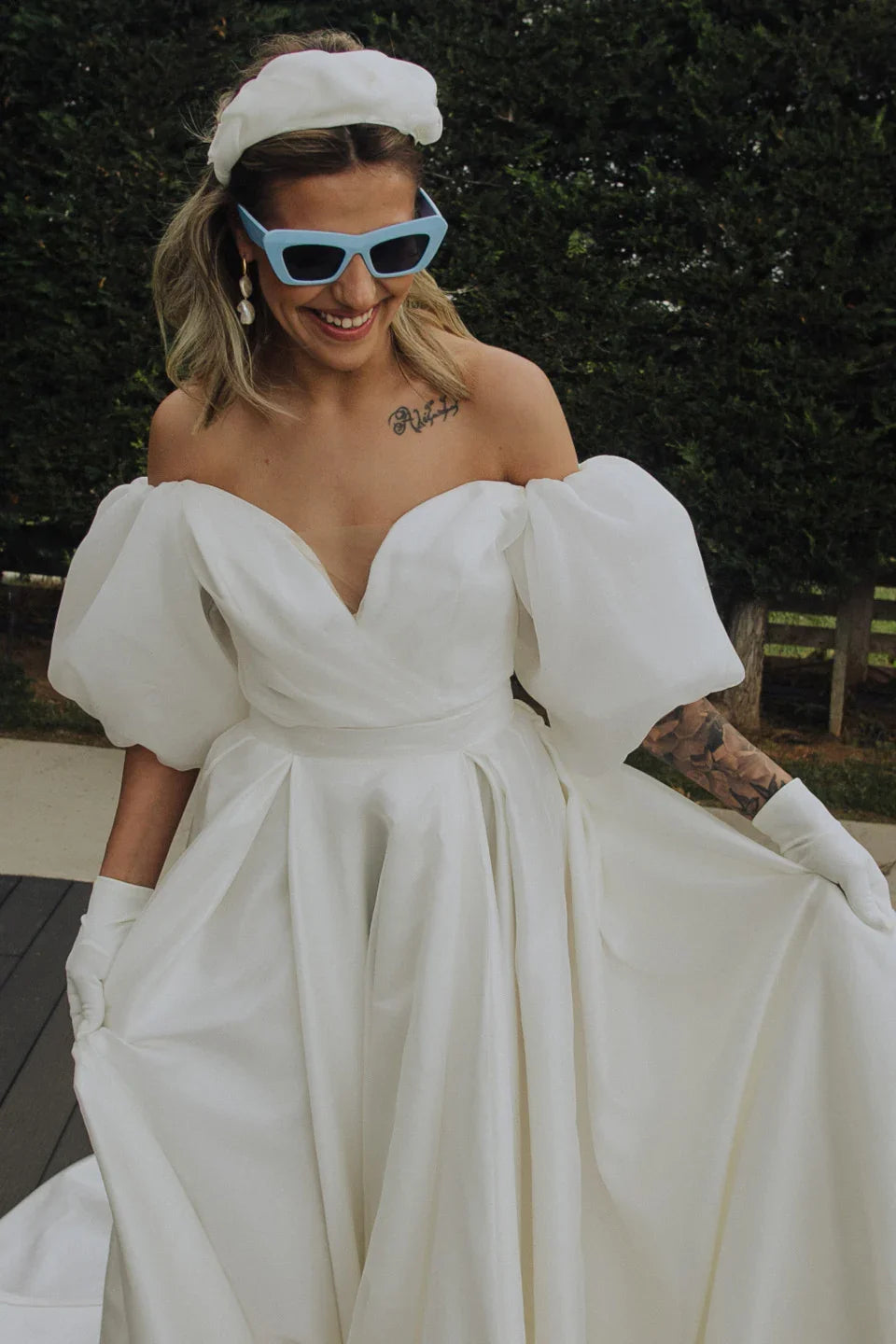 Smiling bride in off-shoulder white wedding dress with puff sleeves, blue sunglasses, white gloves, and headband