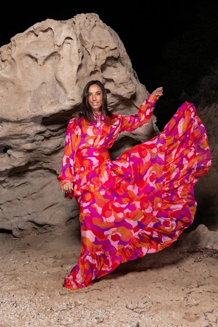 Smiling woman wearing vibrant red, pink, and orange patterned dress posing on sandy ground near large rock formation at night