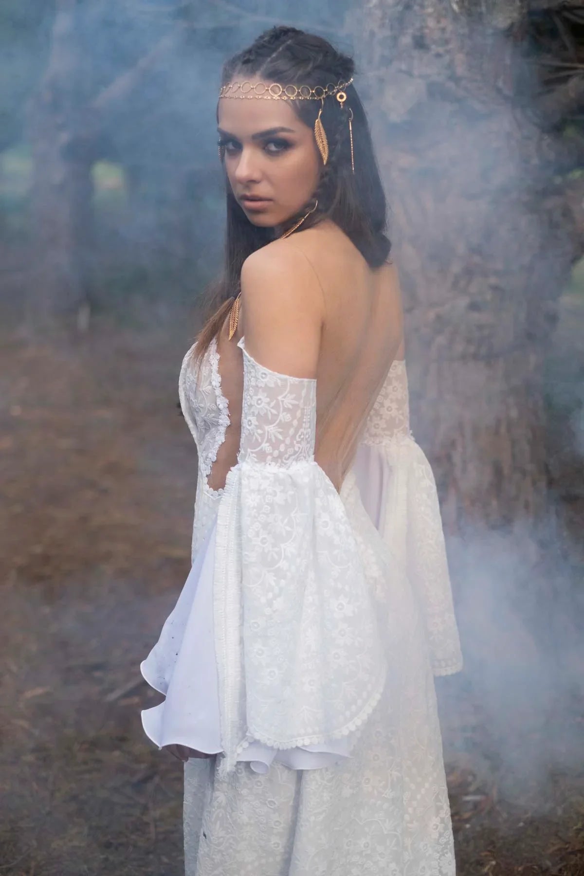 Woman in white floral lace off-shoulder dress with bell sleeves and gold leaf headpiece, smoky forest background