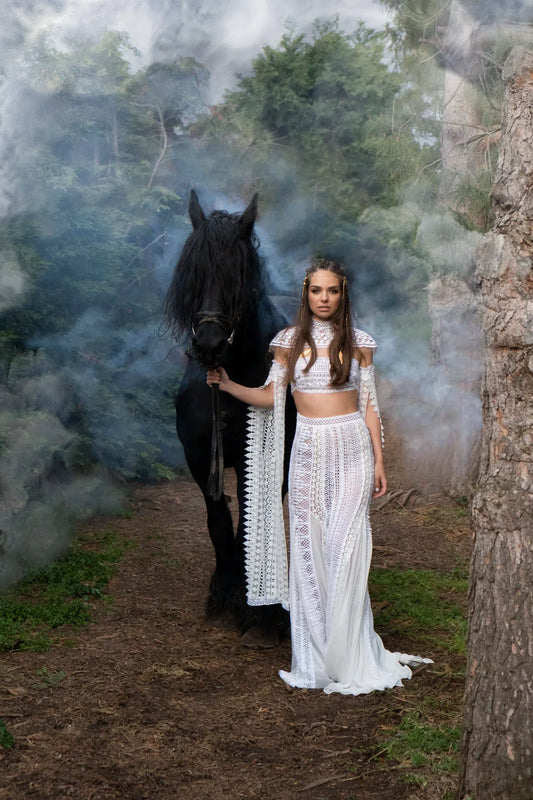 Woman in white lace dress standing beside black horse in smoky forest setting