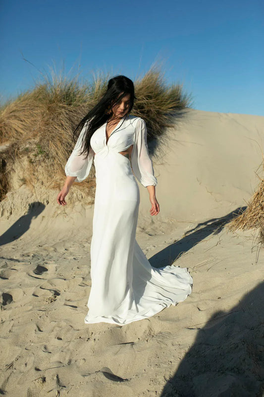 Woman in a flowing white dress with sheer sleeves standing on sandy dunes under a clear blue sky