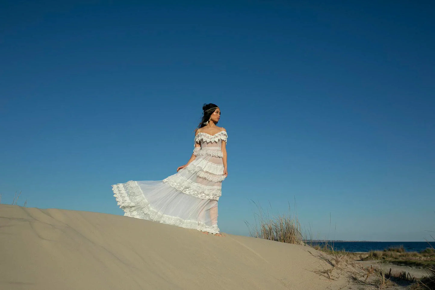 Woman in white lace off-shoulder dress standing on sand dune under clear blue sky near beach