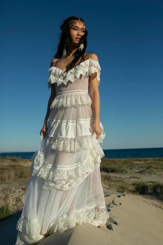 Woman in white lace tiered dress and headchain standing on sandy beach with ocean backdrop