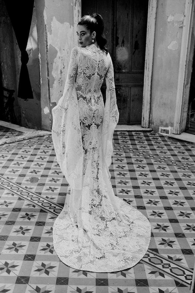 Woman in a long, intricate lace bridal gown with dramatic sleeves, standing on patterned tile floor