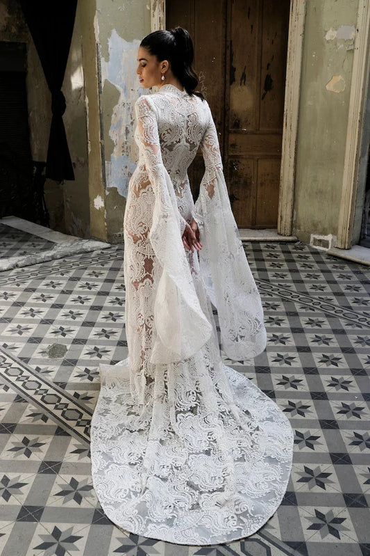Woman wearing a detailed white lace bridal gown with long bell sleeves in rustic room with patterned tiled floor