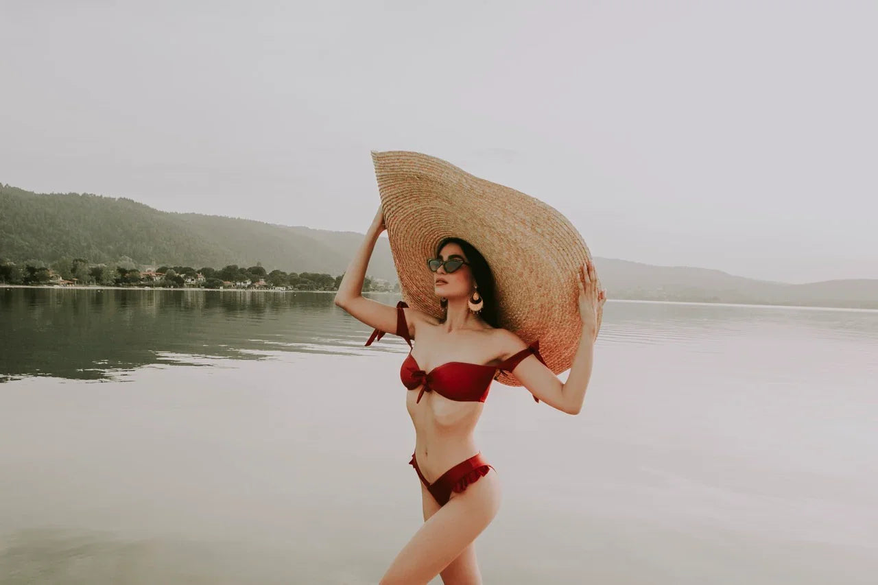 Woman in red bikini and oversized straw hat posing by calm lake with mountains in background