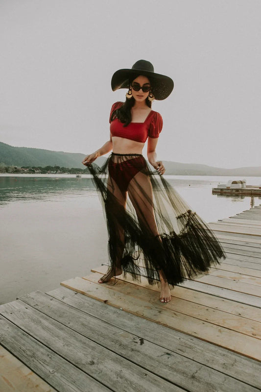 Woman in red bikini and black sheer skirt posing on wooden pier by calm lake at sunset