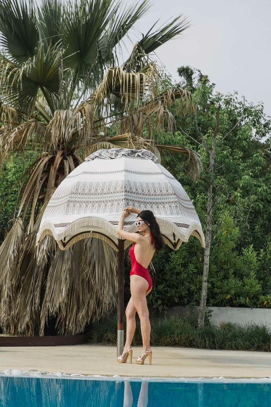 Woman in red swimsuit and heels posing by patterned white umbrella near pool with tropical greenery