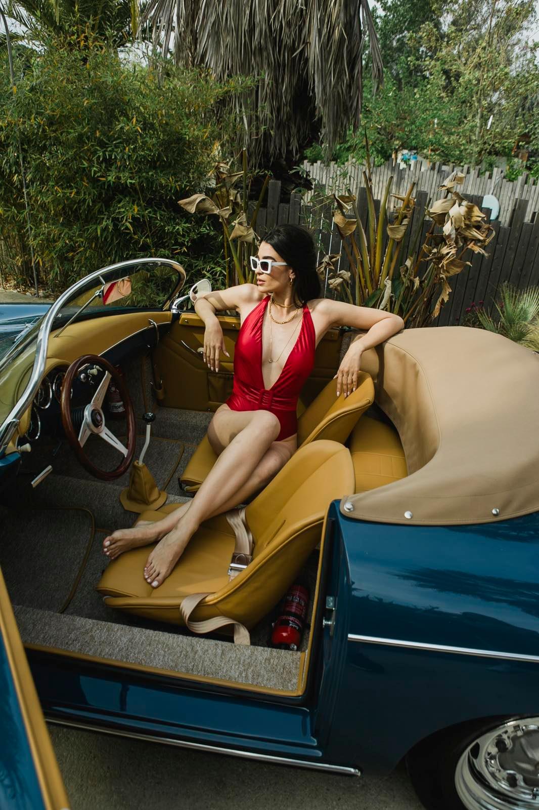 Woman in red swimsuit and white sunglasses relaxing in tan leather seat of vintage blue convertible car with tropical plants background