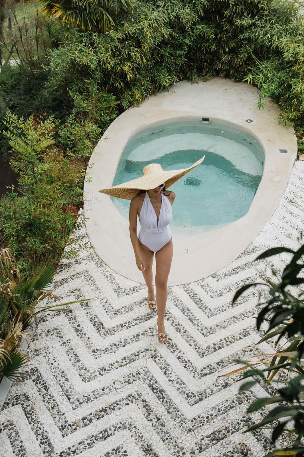 Woman in white swimsuit and oversized straw hat walking by small pool surrounded by lush greenery