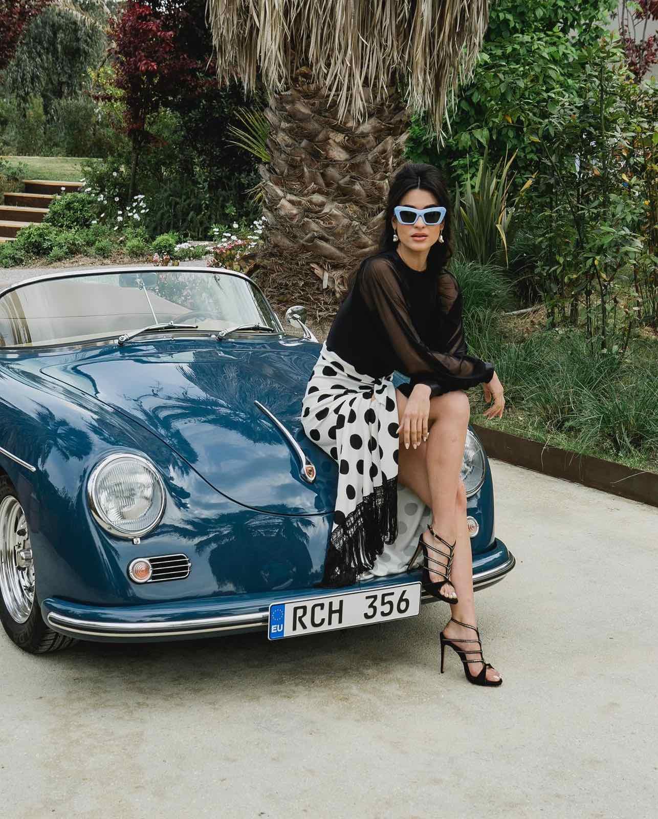 Stylish woman in black and white polka dot skirt sitting on blue vintage convertible car by palm tree