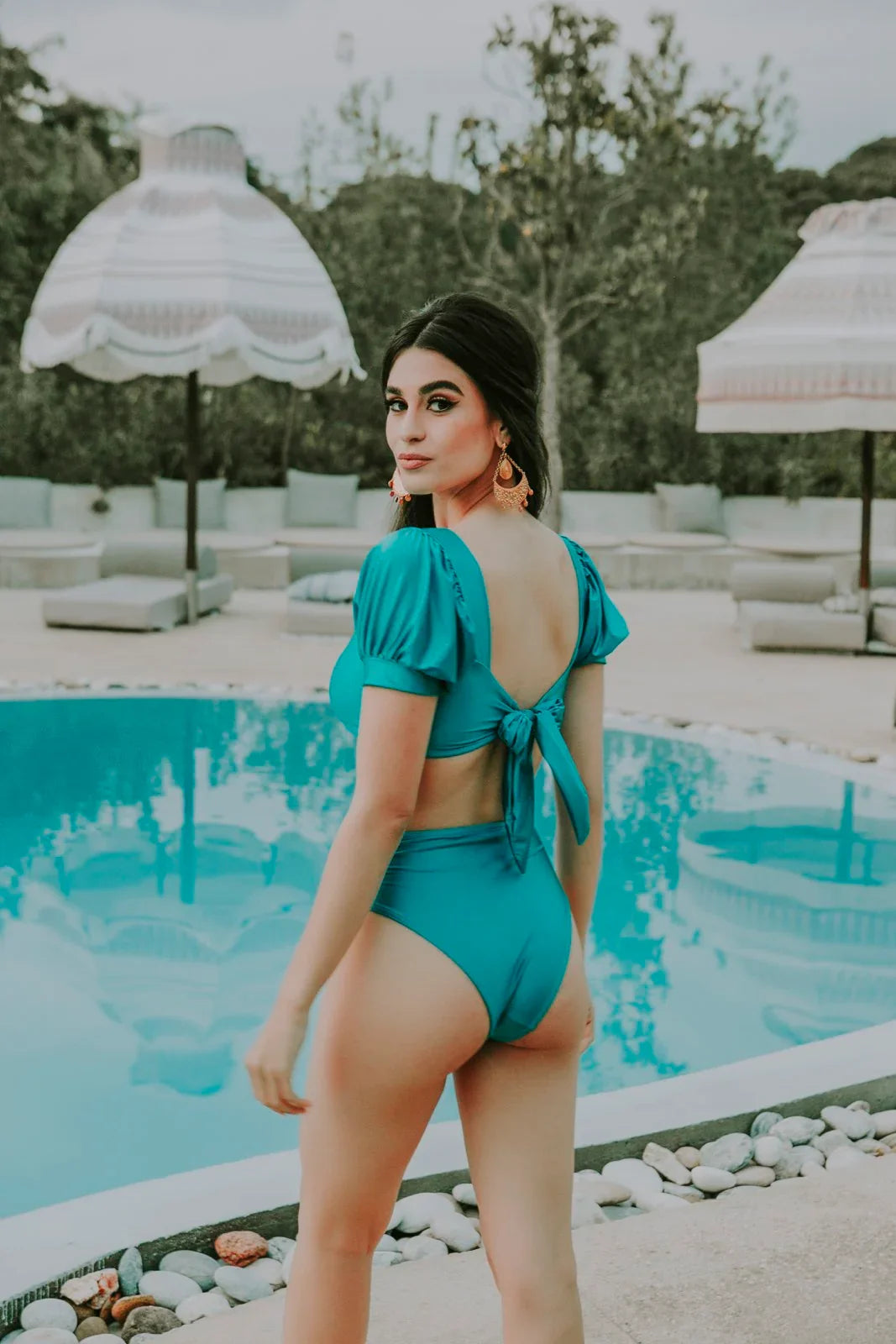 Woman in teal two-piece swimsuit with puff sleeves and back tie near upscale poolside lounge area