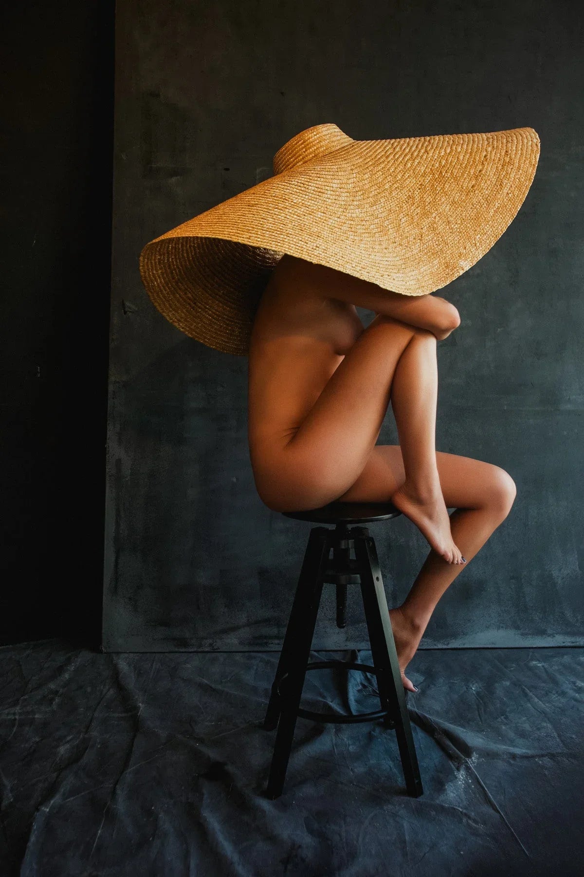 Nude woman sitting on black stool wearing oversized wide-brimmed straw hat against dark backdrop
