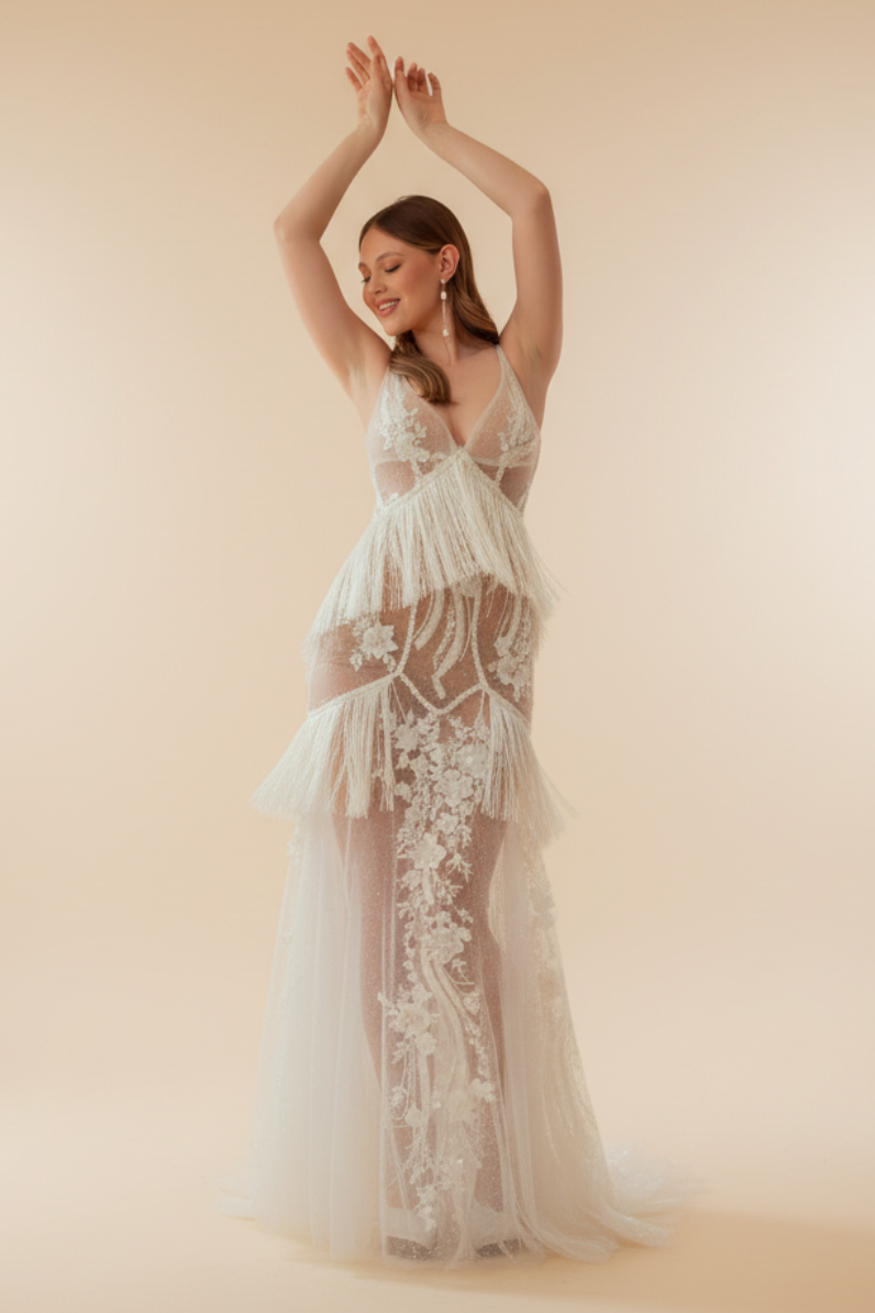 A woman in a white, floral, Maxi dress with a V-neck, standing in a room with a red and beige backdrop, posing with her arms raised.