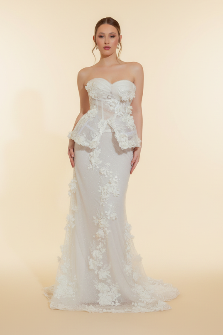 Woman in a white wedding dress standing in a room with patterned wallpaper and wooden floor.