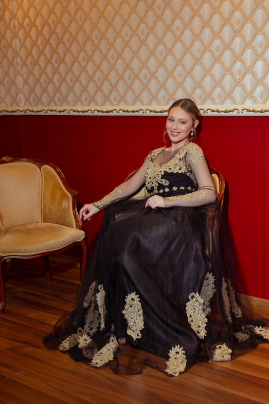 Woman in a black and gold dress sitting on a chair against a red wall with patterned wallpaper.