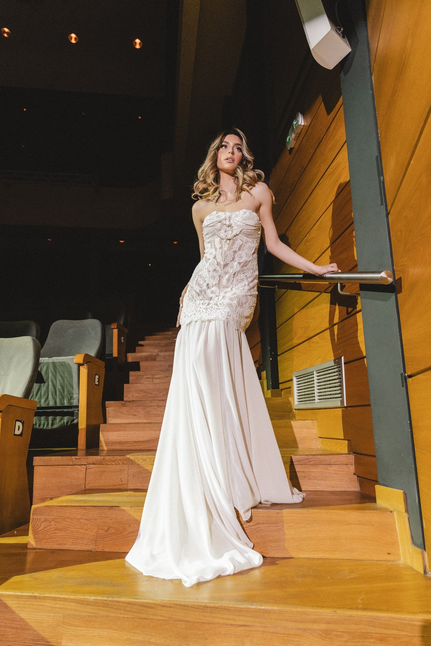 Woman in a white strapless gown standing on wooden stairs inside a building.